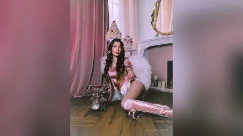Glamorous woman in a pink satin angel costume with white wings sitting on a floor in a dreamy pink room.