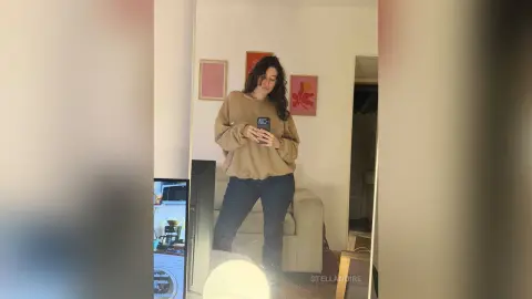 StellaNoire stands in a room wearing a tan sweatshirt and dark pants, holding her phone to take a mirror selfie.