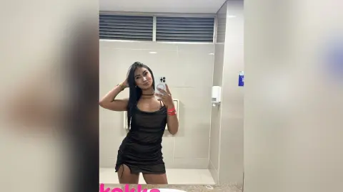 Young woman in a black slip dress taking a mirror selfie in a modern white tiled bathroom.