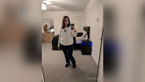 Young woman in a white button-down shirt and dark jeans taking a mirror selfie in a modern bedroom.