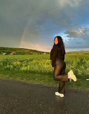 Smiling woman in black top and leather pants standing on a road under a rainbow in a green field.