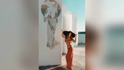ReyQuins poses in a red bikini top and long skirt next to a mermaid mural on a sunny balcony.