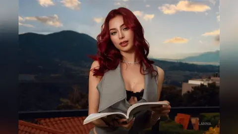 Red-haired woman in a grey sleeveless top reading a book on a balcony overlooking a scenic mountain landscape.