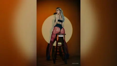 Blonde woman in black lingerie and red lace tights sitting on a stool against a warm sunset background.
