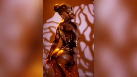 Glamorous Black woman in gold body paint posing against warm patterned shadows in a moody studio setting.