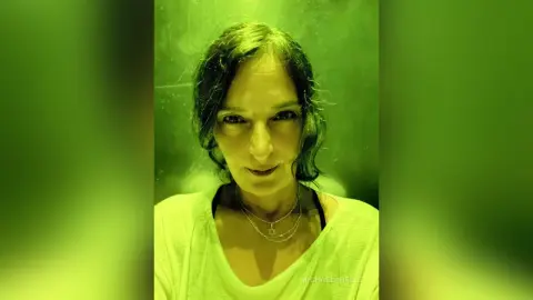MichaelaBelle looks directly at the camera wearing a white v-neck top with her dark hair styled in loose waves.
