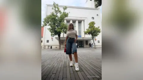 SelenaRusse stands on a paved street in front of a large white building, wearing a tan top and denim shorts.