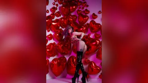 LeyaTaryn poses in a red bodysuit and tall black boots against a background of red heart balloons.