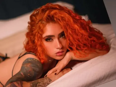 Woman with bright orange curly hair and tattoos relaxing on a white bed in a moody indoor setting.