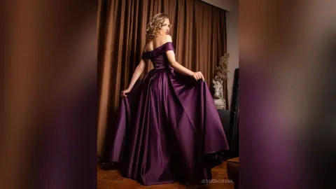 Elegant woman in a flowing purple satin evening gown posing against brown curtains in a moody indoor setting.