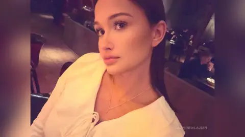 JazminHalajian poses in a white top with a low neckline, showing her long dark hair and neck in a dim indoor setting.