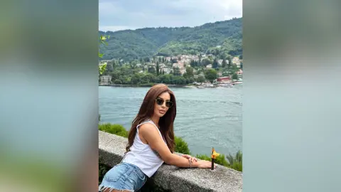 ReynaGomes poses outdoors by a lake, wearing a white tank top, denim shorts, and sunglasses with her long brown hair down.