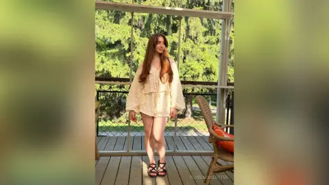 Young woman in a cream lace bohemian dress standing on a sunny wooden balcony surrounded by lush green trees.