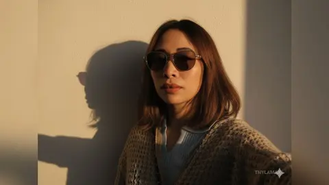 Woman in a cozy knit cardigan and sunglasses posing against a wall in warm sunset lighting.