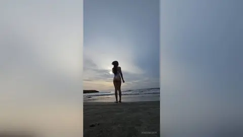RossiNoleen stands naked on a sandy beach at sunset, showing her bare back and profile against the ocean waves.
