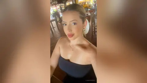 Smiling young woman in a black strapless top and gold jewelry posing in a sunlit outdoor cafe setting.