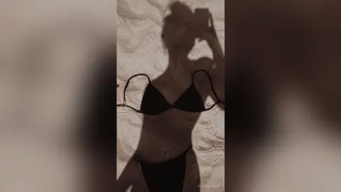 MarisaGrace poses in a black bikini top and bottoms against a sandy beach background, holding a phone up to her face in a shadow-heavy silhouette pose.
