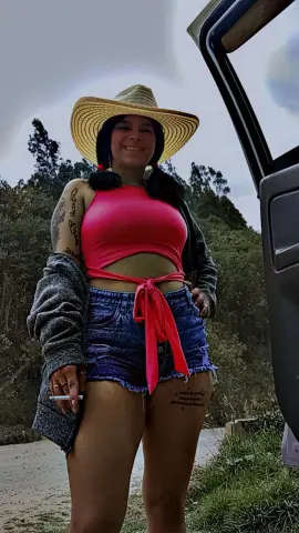 Smiling woman in a pink crop top, denim shorts, and straw hat posing outdoors by a vehicle.