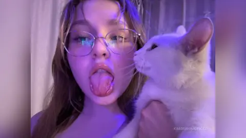 Young woman with glasses playfully sticking out her tongue while cuddling a white cat in purple ambient lighting.