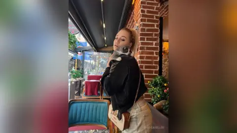 Blonde woman in a black sweater holding a grey kitten outside a brick cafe on a sunny day.