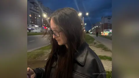 LizbethLocantore wears glasses and a black leather jacket while standing outside on a street at dusk or night.