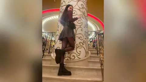 Young woman in a dark sweater and plaid skirt leaning against a decorative pillar in a luxury hotel lobby.