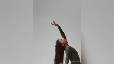 Graceful woman in a dark long-sleeve top stretching her arm against a minimalist grey background.