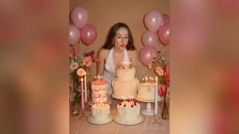 Woman in white dress celebrating with pink balloons and multiple white birthday cakes in a dreamy aesthetic setting.