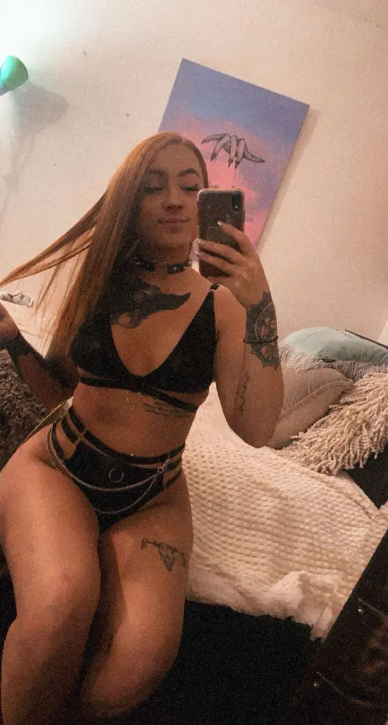 Smiling woman with black bangs and tattoos wearing a red off-the-shoulder top in a cozy bedroom setting.