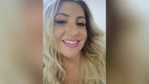 Smiling blonde woman with glamorous makeup posing for a bright, cheerful selfie indoors.