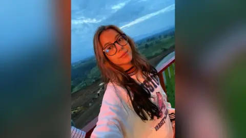 JessieLiu wears glasses and a white graphic sweatshirt while posing outdoors against a blue sky and green landscape.