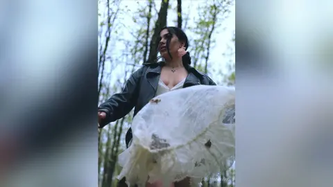 Moody portrait of a woman in a black leather jacket and white lace dress standing in a lush green forest.