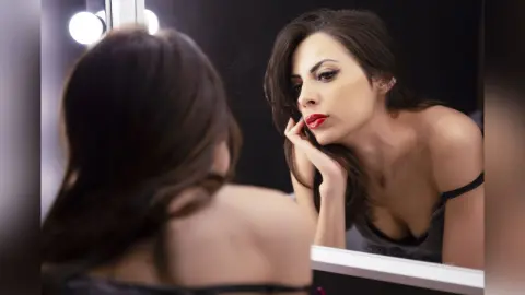 OliviaLarson looks into a mirror with her hand under her chin, wearing a dark grey camisole and red lipstick.