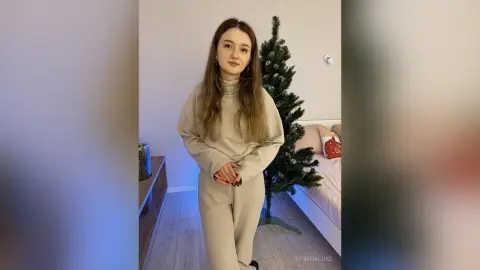 Young woman in a beige lounge set standing in a cozy minimalist bedroom next to a Christmas tree.