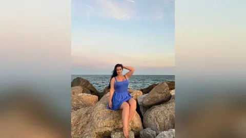 JasminNise poses sitting on large rocks by the ocean, wearing a short blue sundress with her hand behind her head.