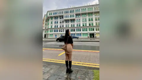 Young woman in a black top and beige skirt standing on a city street in front of a large apartment building.
