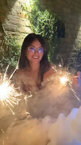 Smiling woman in glasses holding glowing sparklers against a lush green garden wall at night.
