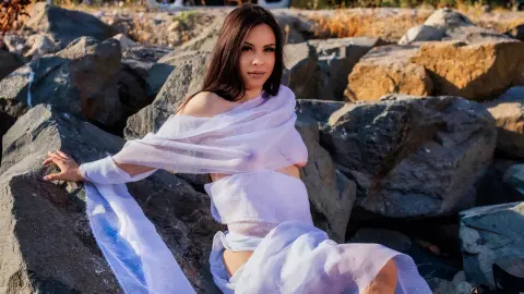 Young woman in a flowing white sheer wrap sitting on large rocks in warm sunlight.