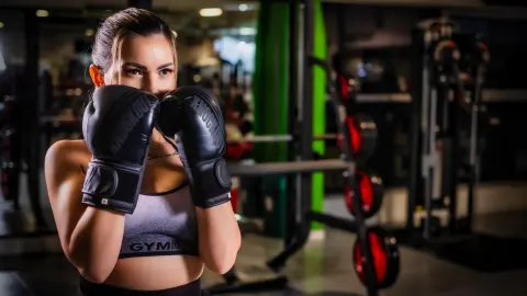 AmiraBelle poses in a gym wearing black boxing gloves and a grey sports bra, looking directly at the camera with her hair pulled back.