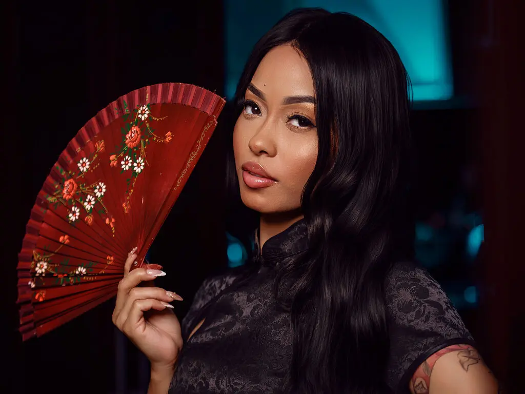 Elegant woman in a black floral qipao holding a red decorative folding fan in a moody, dim setting.