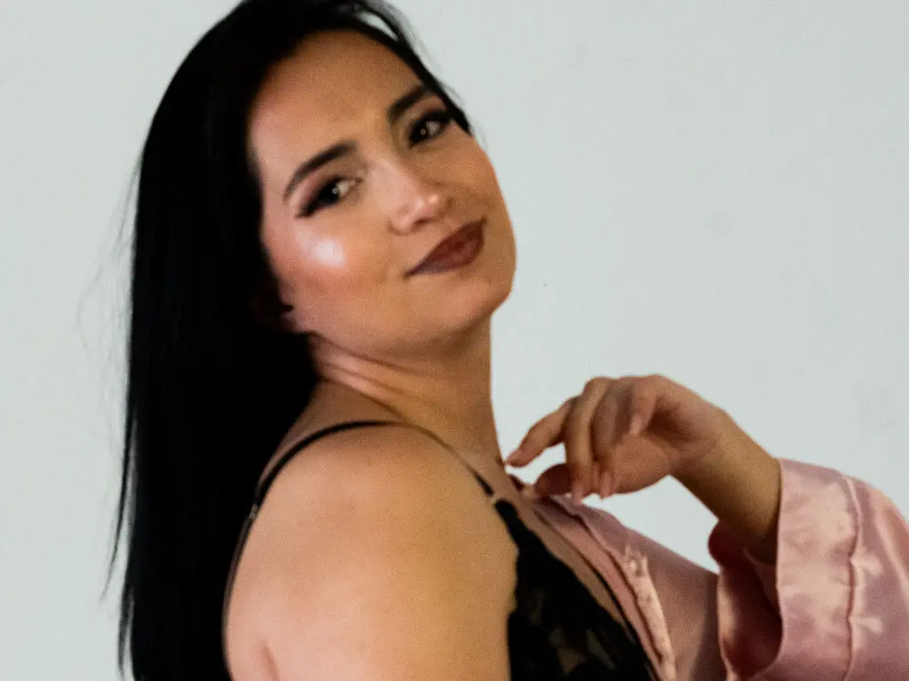Smiling woman with long dark hair wearing a black lace camisole and pink robe in a bright studio setting.