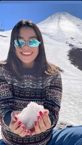 Smiling woman in a patterned sweater holding snow against a sunny mountain landscape.