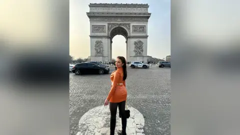 Stylish woman in an orange blazer and black leggings posing in front of the Arc de Triomphe in Paris.