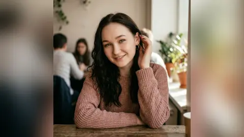 EllaVietor smiles at the camera while sitting at a cafe table wearing a pink ribbed sweater.