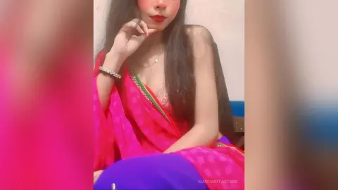 Young woman in a vibrant pink and green traditional saree sitting gracefully in a soft indoor setting.