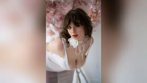 Dreamy portrait of a woman in a white lace camisole with a floral choker in a soft pink floral setting.