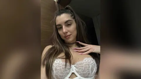 Young woman with long brown hair wearing white lace lingerie in a moody indoor setting.