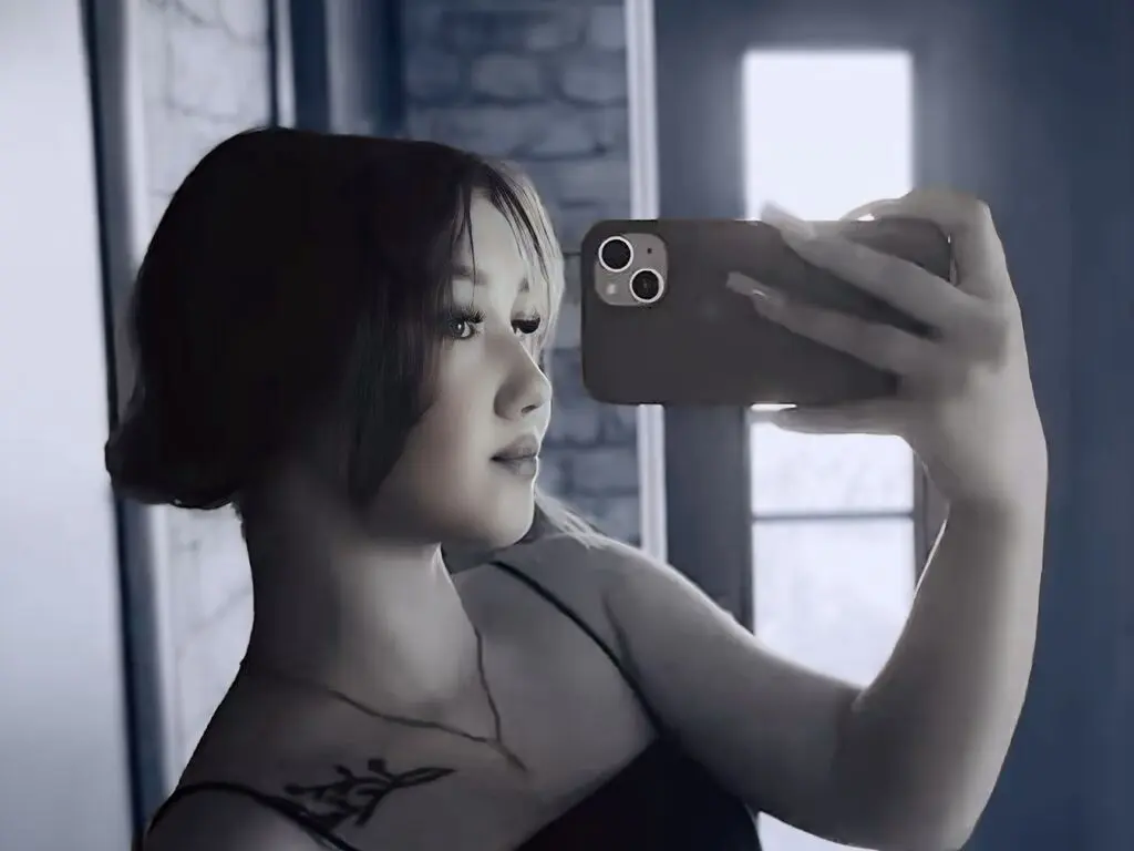 Young woman in a black tank top taking a moody selfie in a mirror near a window.