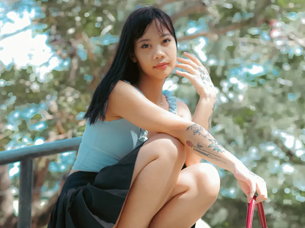 Young woman in a light blue tank top and black skirt posing outdoors against a lush green leafy background.