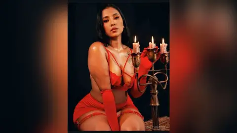 Glamorous woman in red lingerie and gloves posing by lit candles in a moody, dark setting.